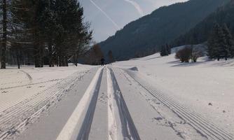 A snowy landscape with cross-country skiing tracks in the snow. In the background, there are wooded hills and a blue sky.