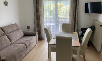 A modern living room with a sofa bed and a dining table. The room features large windows that let in plenty of daylight.