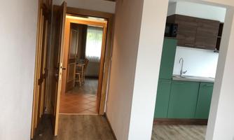 A bright hallway with two open doors leading to different rooms. On the left is a kitchen with green cabinets and a window.