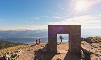 A view of a mountainous viewpoint with a monumental structure in the foreground. Two people are standing in the background and enjoying the view.