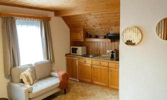 A cozy room with a gray sofa and a wooden kitchen. The room has a warm atmosphere due to the wooden walls and natural light.