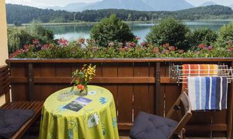 A cozy balcony with a round table and two chairs. In the background, there are mountains and a sparkling lake visible.