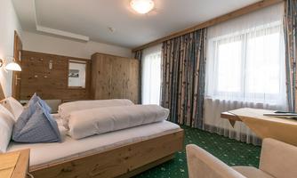 A cozy bedroom with a large bed, wooden furniture, and a table. Large windows provide plenty of natural light and a friendly atmosphere.