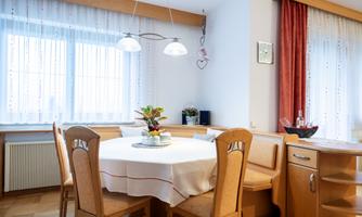 A cozy dining area with a round table and chairs. The windows are adorned with light curtains, and a fruit table decoration gives the room an inviting atmosphere.