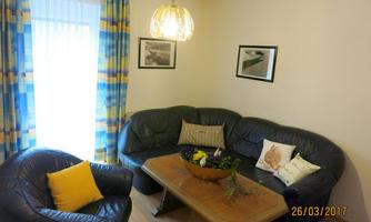 A cozy living room with a blue leather couch and a wooden table. There are decorative pillows and colorful curtains that let light in.