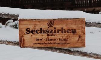 A wooden sign with the inscription "Sechszirben" stands in the snow. In the background, a rustic building can be seen.