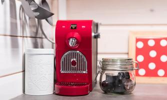 A red coffee machine sits on the kitchen counter next to a white container and a glass with small black capsules. In the background, there is a decorative box with red dots.