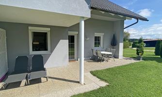 A relaxed terrace with two loungers and a table outdoors. In the background, there is a well-maintained garden and a blue sky.