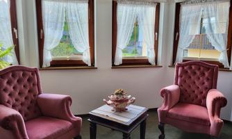 A cozy room with two pink armchairs and a table in the middle. Bright curtains hang in front of the windows, and the view outside is visible.