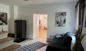 A modern living room with a couch and a rug. In the background, a bright room with decorative elements can be seen.