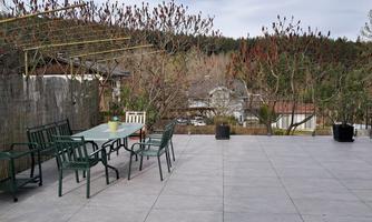 A spacious outdoor area with a table and several chairs. In the background, there are trees and a wooded slope.
