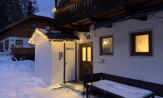 A winter scene with snow-covered ground and festive lighting on a cozy building. The warm glow of the windows creates an inviting atmosphere in the twilight.