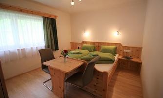 A cozy room with a double bed and a wooden table. The walls are light and the window has a curtain in dark green.