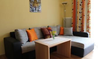 A cozy living room with a gray sofa and colorful cushions. On a wooden table, there is a small vase with fresh flowers.