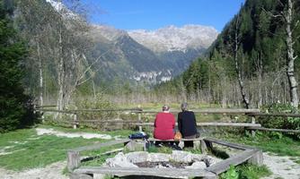 Two people are sitting on a bench and looking at the mountains. The landscape is green with trees and a clear blue sky.