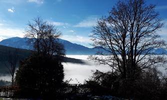 A picturesque landscape with mountains and fog drifting over the valley. In the foreground, bare trees stand, surrounded by gentle hills.