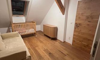 A cozy room with wooden floors and a wooden ceiling. In the room, there is a sofa, a crib, and a small wardrobe.