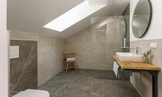 A modern bathroom with gray tiles and plenty of natural light. There is a shower, a sink on a wooden table, and a small seating area.