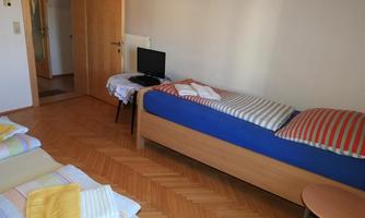 A simple room with two beds and a small table. A television is on a chair, and the walls are painted light.