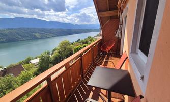 A balcony overlooking a quiet lake and green hills. There is a small table and a hanging chair on the balcony.