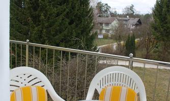 A balcony with two chairs and a table. In the background, green trees and a few buildings can be seen.