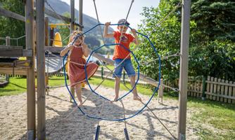 Two children are playing on a climbing frame outdoors. In the background, there are trees and a gentle hill.