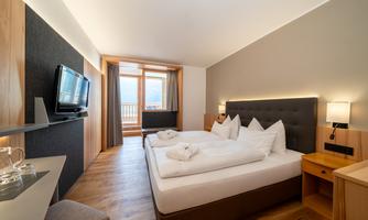 A modern bedroom with a large bed and white pillows. Large windows offer a view of nature and provide plenty of daylight.
