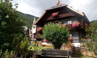 A traditional wooden house with a green garden and colorful flowers. In the background, there is a church tower and the surrounding mountain landscape.