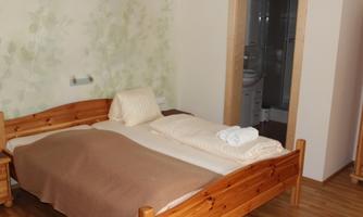 A simply furnished bedroom with a wooden double bed and neutral colors. A bathroom is visible in the background.