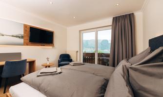 A modern room with a double bed, a television, and a comfortable armchair. Through the windows, there is a beautiful view of the mountains.