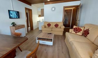 A cozy living room with a beige sofa and red cushions. A dining table and a modern kitchen are also visible.