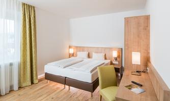 A modern room with a double bed and a desk. The walls are bright, and large windows provide plenty of daylight.