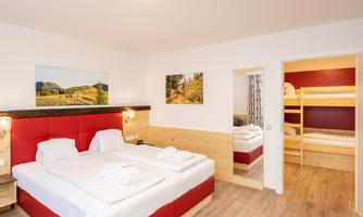 A modern hotel room with a double bed and a sleeping area for children. The walls are done in light wood, and there are pictures of landscapes.