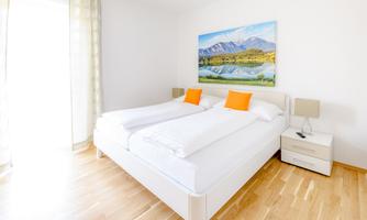 A bright bedroom with a large bed and orange pillows. A beautiful landscape painting with mountains and a lake hangs on the wall.