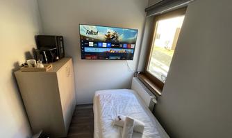 A small room with a single bed and a television on the wall. Next to the bed is a wooden piece of furniture with a kettle and a coffee machine.