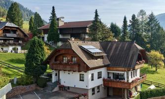The charming wooden house in country house style with parking directly in front of the house. In the background, the green surroundings can be seen.