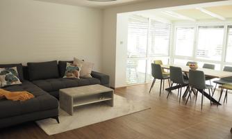 A modern living room with a comfortable sofa and a coffee table. In the background, a bright dining area can be seen with large windows and a dining table.