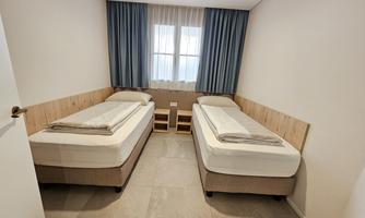 A simple room with two single beds and a window. The walls are light, and there are curtains in a shade of blue.