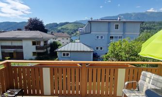 A view from a balcony of an idyllic village with a mountain backdrop. In the foreground, there are a wooden railing, a bench, and a sunshade.