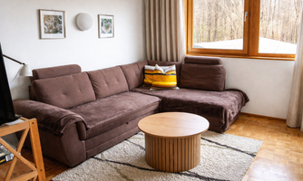 A cozy living room with a brown sofa and a round wooden table. Large windows provide plenty of daylight and a view of the greenery.