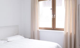 A bright bedroom with a bed and a large window. The curtains are beige and the floor is made of wood.