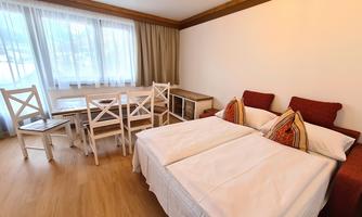 A cozy room with two single beds and a small dining area. Large windows provide plenty of daylight.