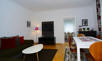 A modern living room with a cozy sofa and a dining table. The walls are light and there are decorative elements like plants and pictures.