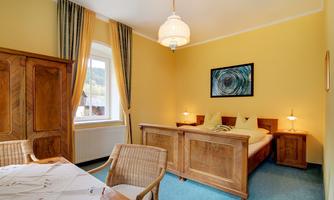 A bright and cozy bedroom with yellow walls and a large window. The room is furnished with a wooden wardrobe and a table, as well as comfortable beds.