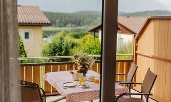 A balcony with a covered table and a view of nature. On the table are breakfast dishes and a flower vase.