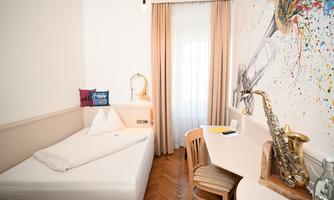 A cozy room with a single bed and a desk. On the wall, there is a colorful design featuring a trumpet.