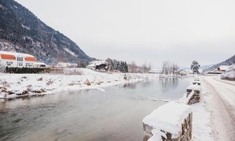 A snowy landscape with a calm river and surrounding mountains. In the background, some houses and trees can be seen.