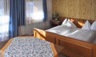 A cozy bedroom with two beds and a table. Large windows let in plenty of light and the walls are made of bright wood.