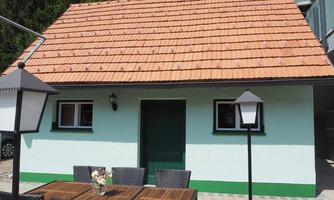 A small, charming house with a red tile roof and a light green facade. Outside, there is a table with chairs, and there are lamp-like lights.