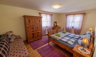 A cozy bedroom with a large bed, wooden furniture, and a sofa. The windows are covered with curtains, and the room feels bright and inviting.